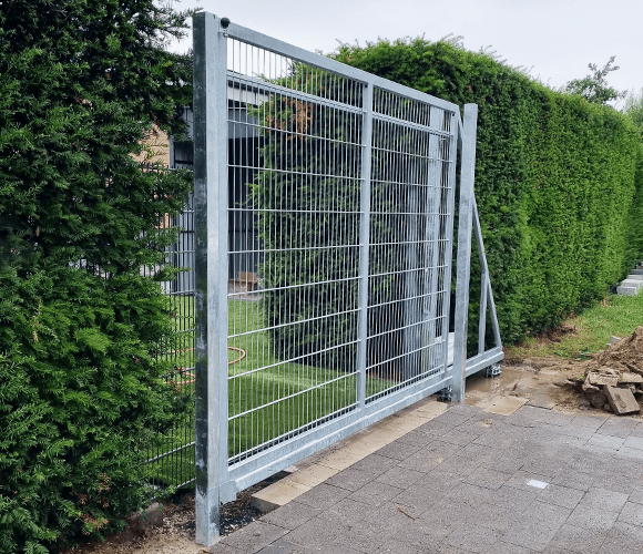 Freitragendes Schiebetor aus verzinktem Stahl mit Doppelstabmatten-Füllung in einem Garten neben einer Hecke.