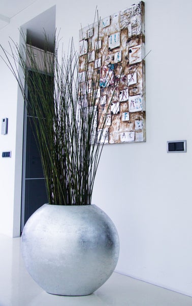 Großer kugelförmiger Pflanzkübel mit hohen Gräsern in einem modernen, hellen Flur neben einem Wandbild und Lichtschaltern.