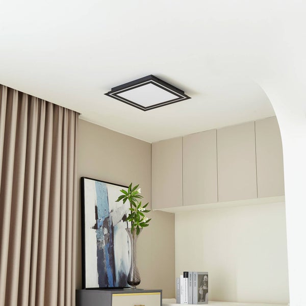 Moderne quadratische LED-Deckenleuchte mit schwarzem Rahmen in einem hellen, minimalistischen Raum mit beigen Vorhängen und Dekoration.