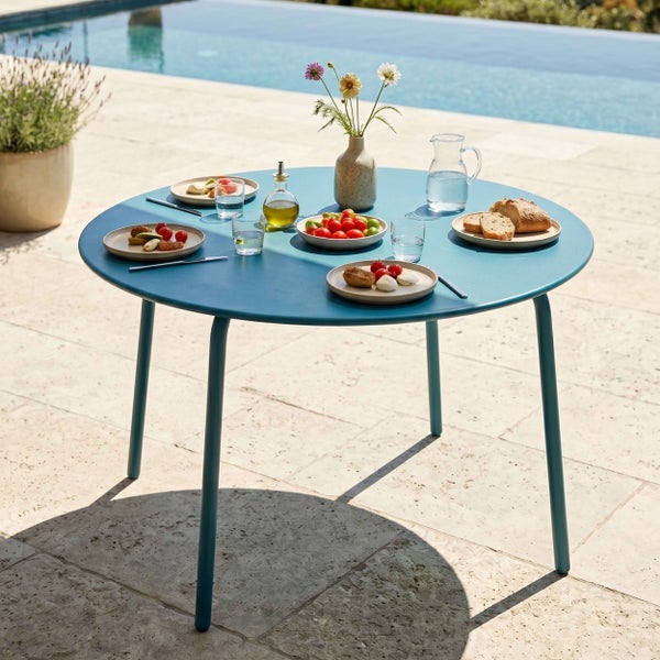 Runder Garten-Esstisch aus Metall auf einer hellen Steinterrasse am Pool, gedeckt mit Speisen, Wassergläsern und einer Blumenvase.