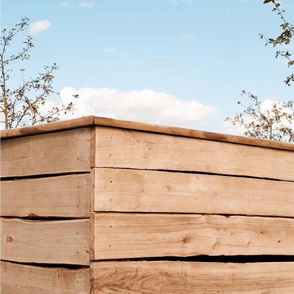 Ein Komposter aus horizontalen Holzbohlen im Garten unter blauem Himmel.