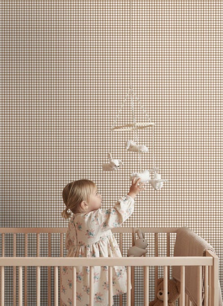 Kinderzimmer mit hell karierter Tapete und Holzgitterbett. Ein Kleinkind greift nach einem Mobile mit Stofftieren.