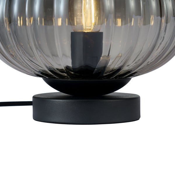 Detailansicht einer Tischleuchte mit rundem schwarzem Metallsockel und geriffeltem Rauchglas Schirm mit leuchtendem Leuchtmittel.