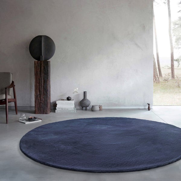 Moderner minimalistischer Raum mit einem großen runden dunkelblauen Hochflor-Teppich auf grauem Betonboden und Dekorationsobjekten.