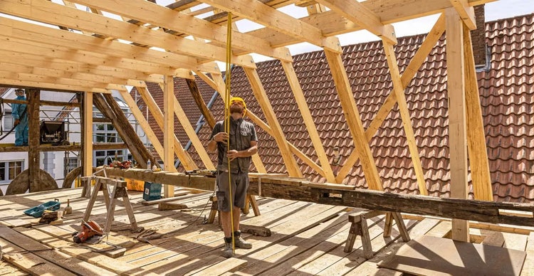 Handwerker misst Holzkonstruktion an einem Dachstuhl im Rohbau mit einem Maßband