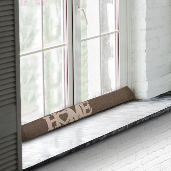 Zugluftstopper mit dem Schriftzug Zuhause auf einer Fensterbank vor einem Fenster