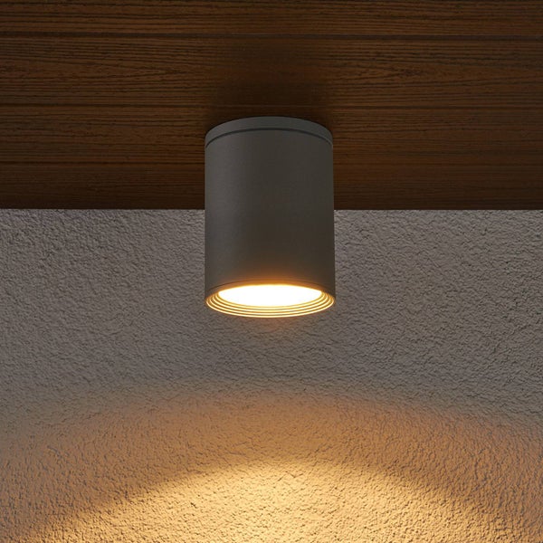 Zylindrische Deckenleuchte aus Metall in Anthrazit, an einer Holzdecke montiert, mit einem warmen Lichtkegel an einer strukturierten Wand.