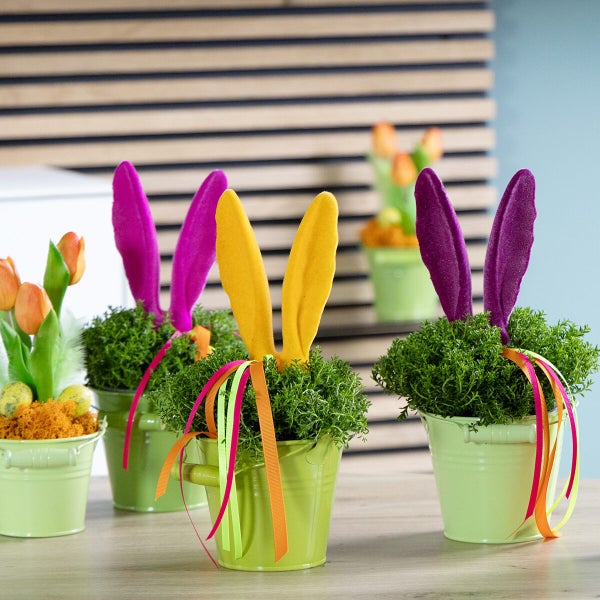 Osterdekoration mit Pflanztöpfen, Osterhasenohren aus Filz, künstlichen Tulpen und Frühlingsgras auf einem Holztisch.