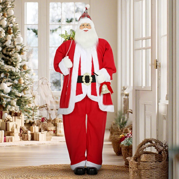Stehende Weihnachtsmann-Figur in rot-weißem Gewand mit karierter Mütze und goldener Glocke in einer hellen weihnachtlich dekorierten Wohnumgebung.