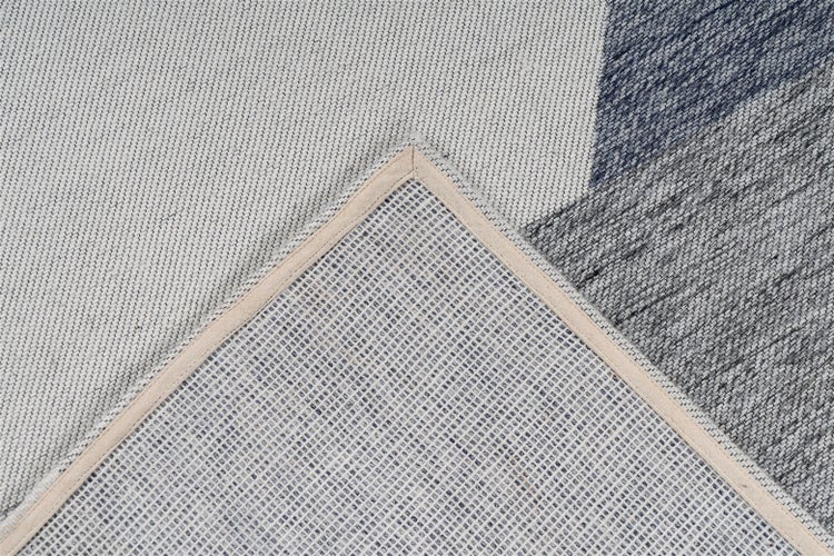 Detailansicht eines gewebten Teppichs mit geometrischem Muster, umgeschlagene Ecke zeigt die gitterartige Rückseite und die verstärkte Kante.