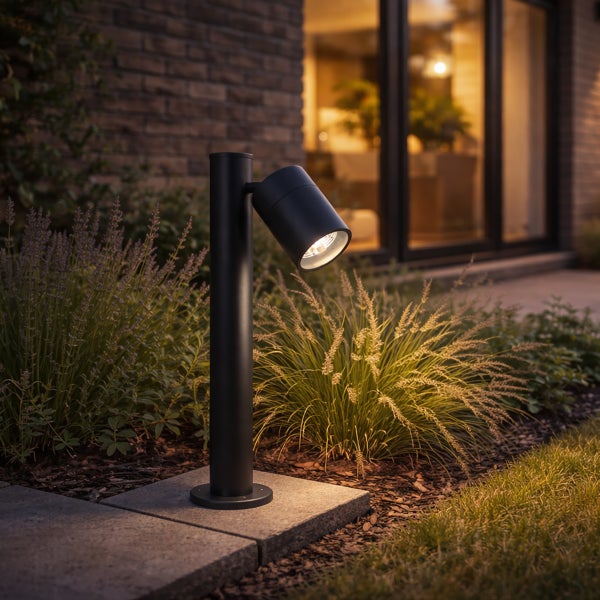 Schwarze LED-Außenleuchte als Gartenstrahler auf einem Steinsockel im Beet neben Gräsern bei Dämmerung.