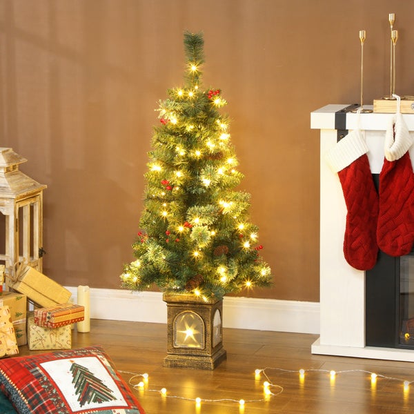 Künstlicher Weihnachtsbaum mit warmweißen LEDs, Beeren und Zapfen in einem beleuchteten Sockel, festlich arrangiert neben einem Kamin.