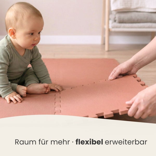Ein Baby sitzt auf einer strukturierten Puzzlematte aus Schaumstoff, während Hände ein neues Teil anfügen. Flexibel erweiterbare Spielmatte.