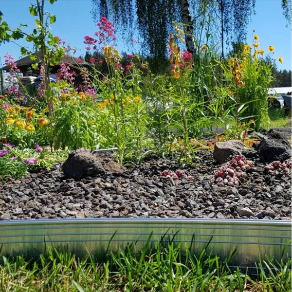 Gartenbeet mit Rasenkante aus Metall, grauem Kies, verschiedenen blühenden Pflanzen und grünem Gras im Vordergrund.