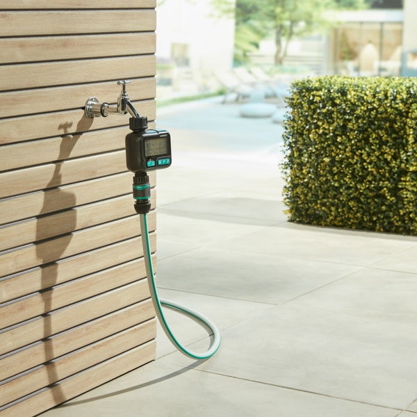 Digitaler Bewässerungscomputer an einem Außenwasserhahn mit angeschlossenem Gartenschlauch vor einer Holzfassade.