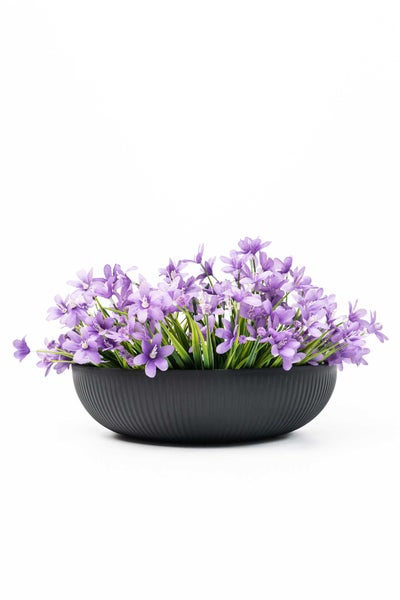 Künstliches Blumenarrangement mit lilafarbenen Blüten in einer runden, dunkelgrauen Schale mit Rillenstruktur.