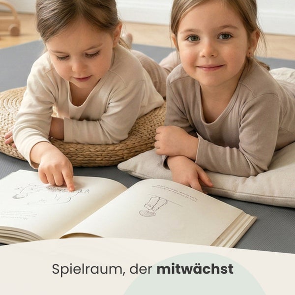 Zwei Kinder liegen in einem hellen Raum auf einer grauen Spielmatte und schauen in ein Buch, gestützt durch ein geflochtenes Sitzkissen und ein Kissen.
