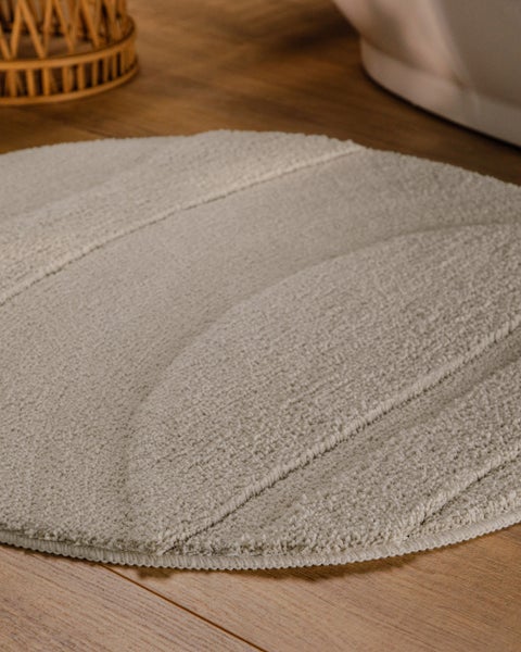 Runder Badeteppich mit strukturierter Oberfläche und Linienmuster auf Holzboden in einem hellen Badezimmer neben einer Badewanne.