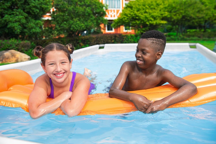 Zwei Kinder in einem rechteckigen Aufstellpool mit weißem Rahmen lehnen auf einer orangefarbenen Luftmatratze in einem sonnigen Garten.