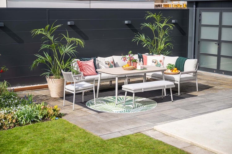 Moderne Garten-Lounge-Ecke mit Ecksofa, Tisch, Bank und Sessel auf einer gepflasterten Terrasse mit dunklem Sichtschutzzaun und Pflanzen.