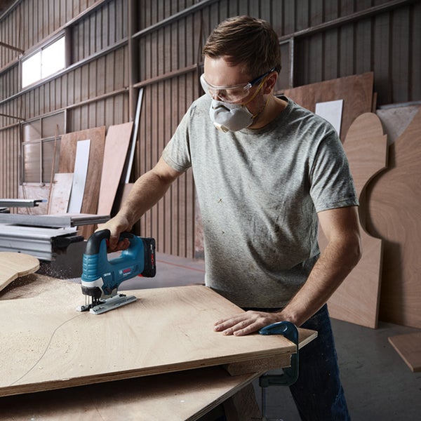 Eine Person mit Schutzbrille und Atemschutzmaske nutzt eine blaue kabellose Stichsäge für Kurvenschnitte in Sperrholz in einer hellen Werkstatt.