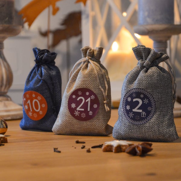 Drei kleine Adventskalender Säckchen aus Jute mit nummerierten Aufklebern für die Weihnachtsdekoration auf einem Holztisch.