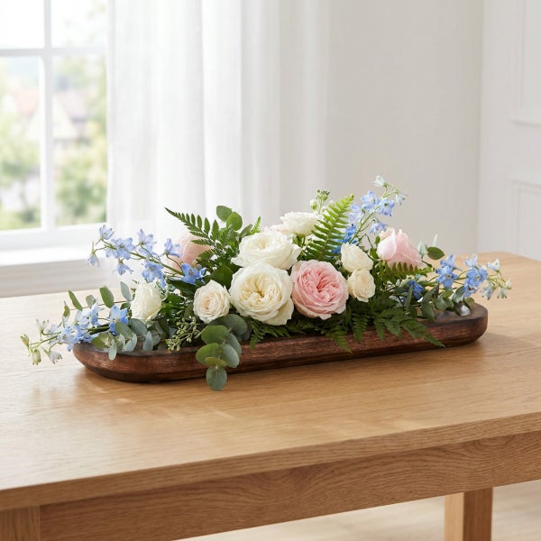 Tischgesteck mit Rosen und kleinen blauen Blüten in einer länglichen Holzschale auf einem Holztisch.