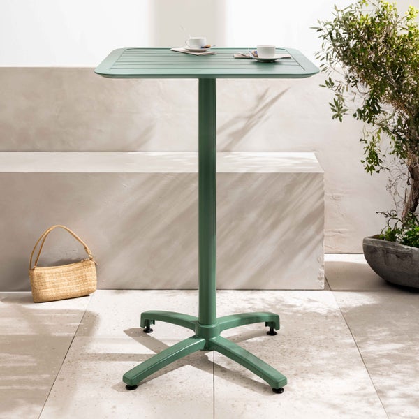 Quadratischer grüner Stehtisch aus Metall mit Lattenstruktur auf einer hellen, organisierten Terrasse mit Steinbank, Pflanze und Strohtasche.