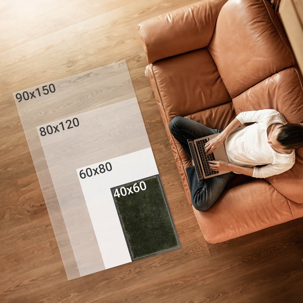 Illustration von Teppichgrößen auf einem Holzboden, mit einer Person auf einem Sofa, die einen Laptop benutzt. Verfügbare Größen sind 90x150, 80x120, 60x80 und 40x60 Zentimeter.