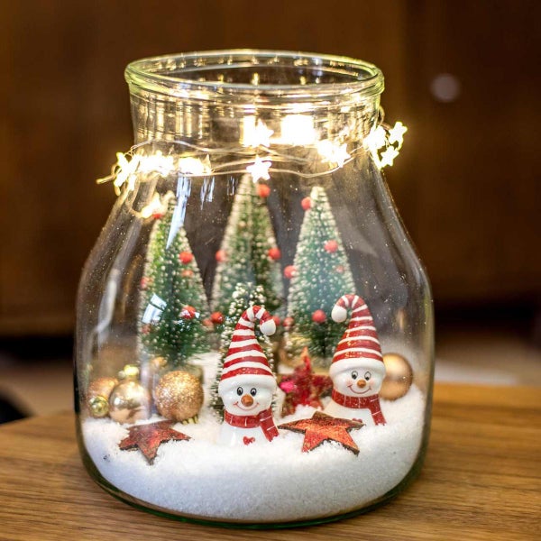 Weihnachtsdekoration im Glas mit Schneemännern, Tannenbäumen, Kunstschnee und LED-Sternen-Lichterkette auf einem Holztisch.