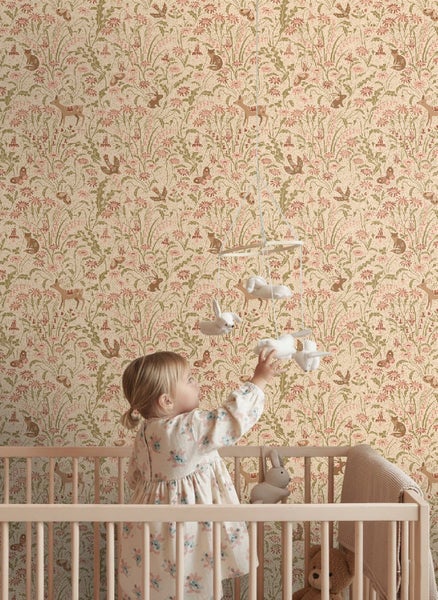 Kleinkind im Gitterbett vor einer Mustertapete mit Waldtieren und Pflanzenmotiven im Kinderzimmer.