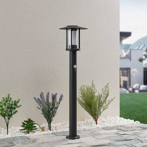 Schwarze Wegeleuchte mit Bewegungsmelder in einem Gartenbeet mit weißen Kieselsteinen und Pflanzen vor einer strukturierten Wand.