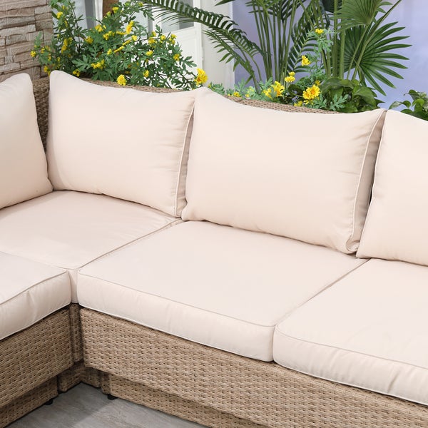 Garten-Ecksofa aus Kunststoffgeflecht mit dicken Polsterkissen vor einer Kulisse aus grünen Pflanzen und gelben Blumen.