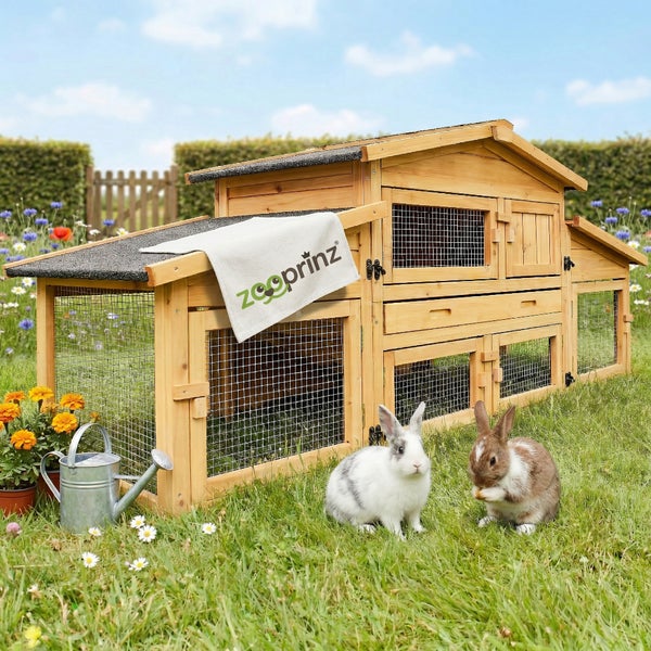 Zooprinz Kaninchenstall aus Holz mit integriertem Freigehege in einem hellen Garten mit zwei Kaninchen, Blumen und Gießkanne.