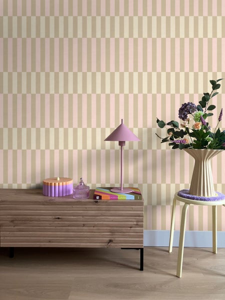 Helles Zimmer mit geometrisch gemusterter Tapete, einem Sideboard aus Holz, einer Tischlampe und einem Blumenarrangement auf einem Hocker.