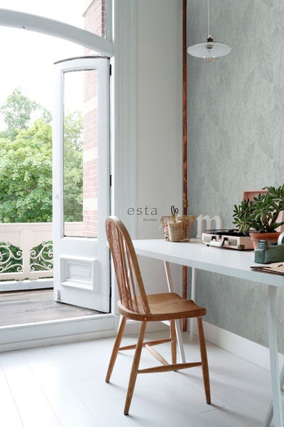Helles Arbeitszimmer mit Vliestapete mit Blättermuster, Esta Home Logo, weißem Schreibtisch, Holzstuhl und offener Balkontür.