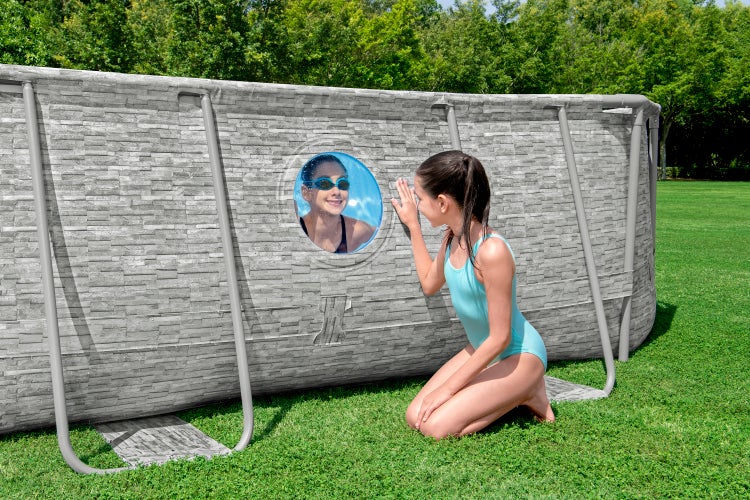 Grauer Aufstellpool in Steinoptik mit rundem Sichtfenster und Metallrahmen im Garten auf einer Rasenfläche.