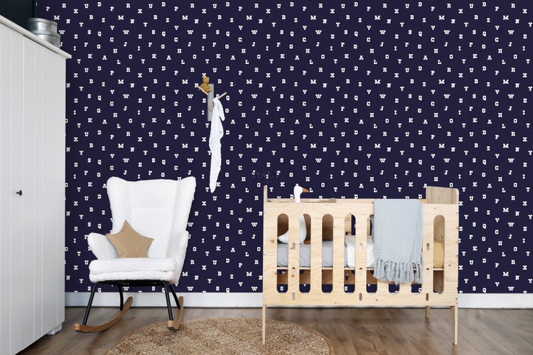 Kinderzimmer mit Gitterbett, Schaukelstuhl und Schrank vor einer Wand mit Buchstabenmuster-Tapete.