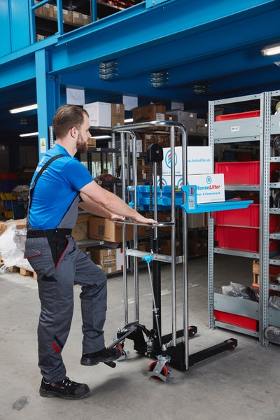 Mitarbeiter in Arbeitskleidung bedient einen manuellen Hydraulikstapler im Lager, um Kartons neben einem Schwerlastregal anzuheben.