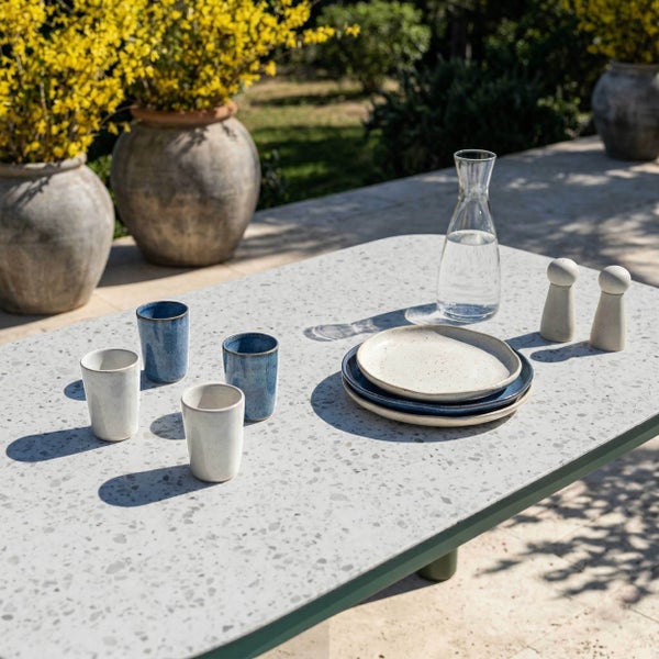Moderner Gartentisch mit Terrazzo-Oberfläche, gedeckt mit Keramikgeschirr und Glaskaraffe auf einer hellen Außenterrasse.