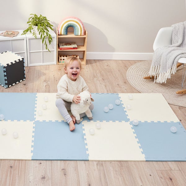 Kleinkind spielt mit einem Stofftier auf einer großflächigen Schaumstoff-Puzzlematte in einem hellen Kinderzimmer mit Holzfußboden.