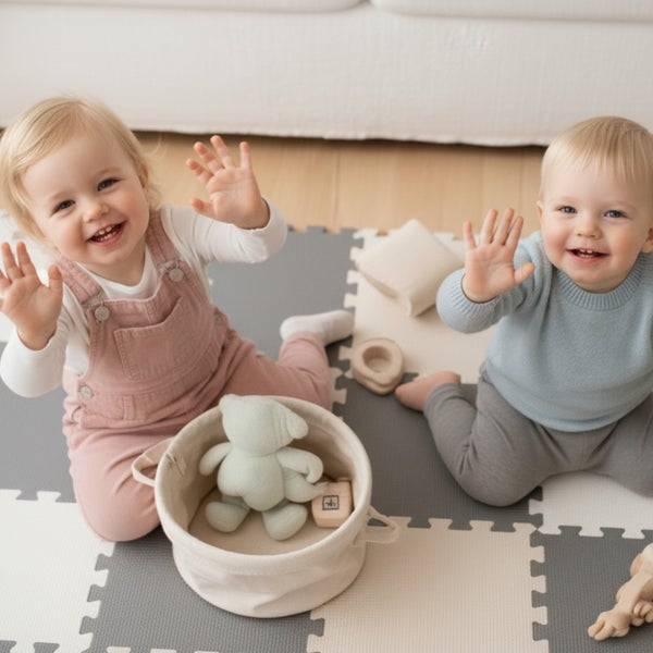 Zwei kleine Kinder spielen auf einer weichen Spielmatte aus Schaumstoff mit Spielzeug und einem Aufbewahrungskorb in einem hellen Kinderzimmer.