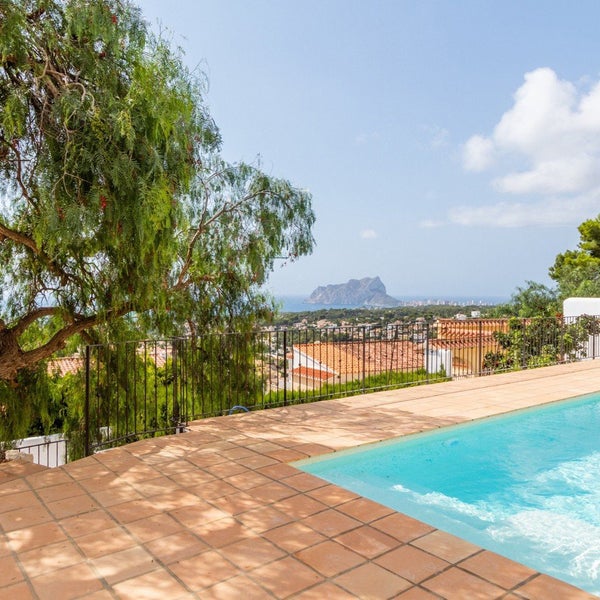 Terrasse mit Fliesenbelag und angrenzendem Swimmingpool mit Blick auf das Meer und eine felsige Küstenlandschaft unter blauem Himmel.