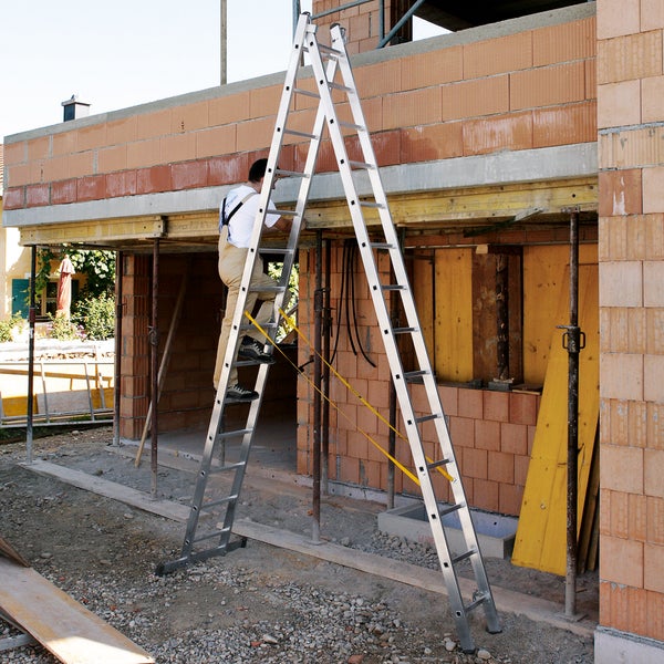 Eine Doppelstufenleiter aus Aluminium steht vor einem im Bau befindlichen Gebäude, auf der ein Bauarbeiter steht.