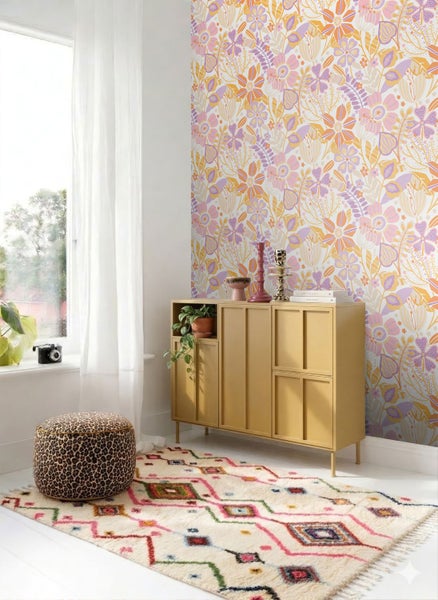 Helles Zimmer mit Vliestapete im Retro-Blumendesign, einem gelben Sideboard aus Holz und einem bunt gemusterten Teppich.