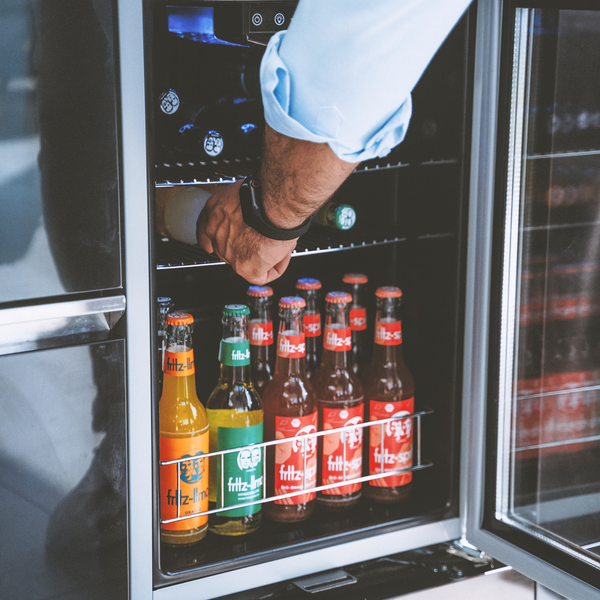 Eine Person greift in einen geöffneten Getränkekühlschrank, der mit verschiedenen Glasflaschen gefüllt ist.