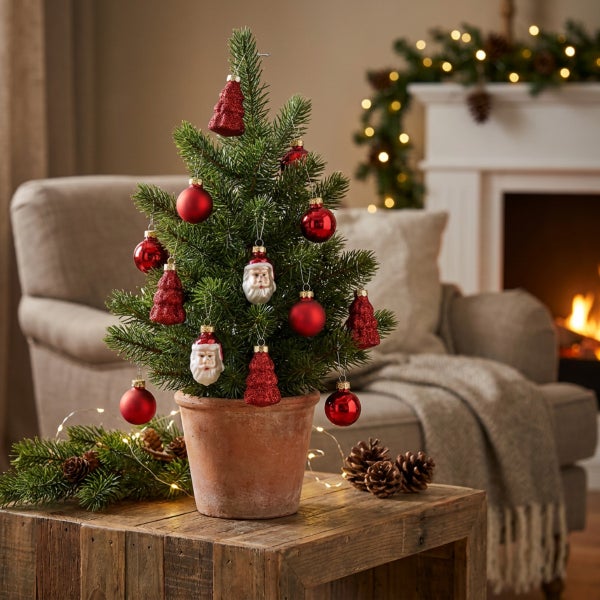 Ein geschmückter künstlicher Weihnachtsbaum in einem Terrakotta Topf auf einem Holztisch, im Hintergrund ein gemütliches Wohnzimmer mit Kamin.