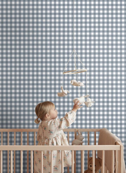 Kinderzimmer mit blau-weiß karierter Tapete, Kind in einem Holzgitterbett greift nach einem Mobile.