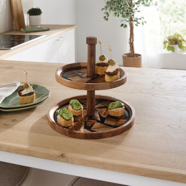 Zweistöckige Etagere aus Holz mit geometrischem Intarsienmuster auf einem Holztisch, belegt mit Häppchen in einer hellen, organisierten Küche.