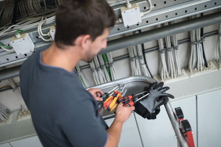 Elektriker hält auf einer Leiter Werkzeuge und Arbeitshandschuhe vor einem Schaltschrank mit zahlreichen Kabeln.